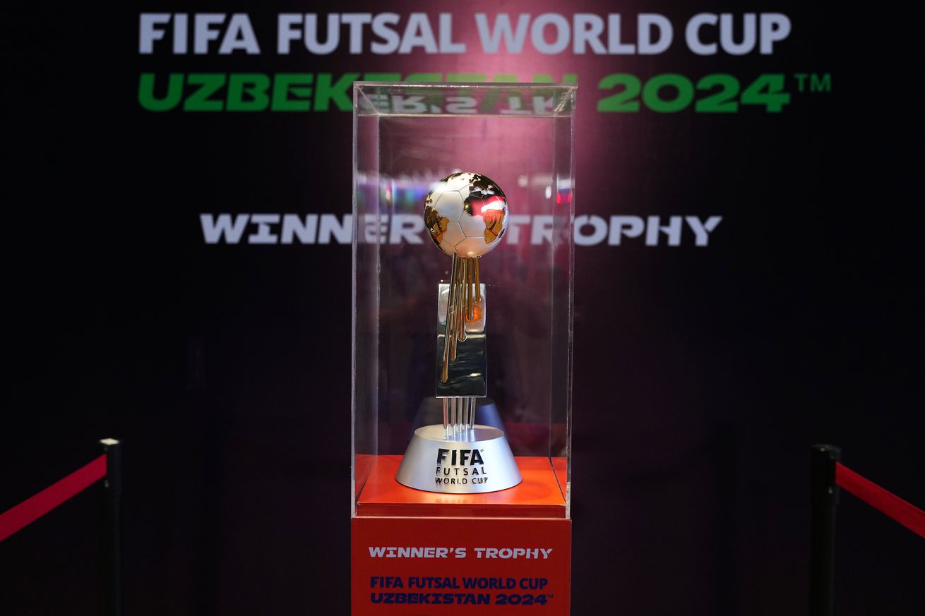 A trophy with a soccer ball on top in a glass case. Behind is a background informing us that it is the Fifa Futsal World Cup winner trophy