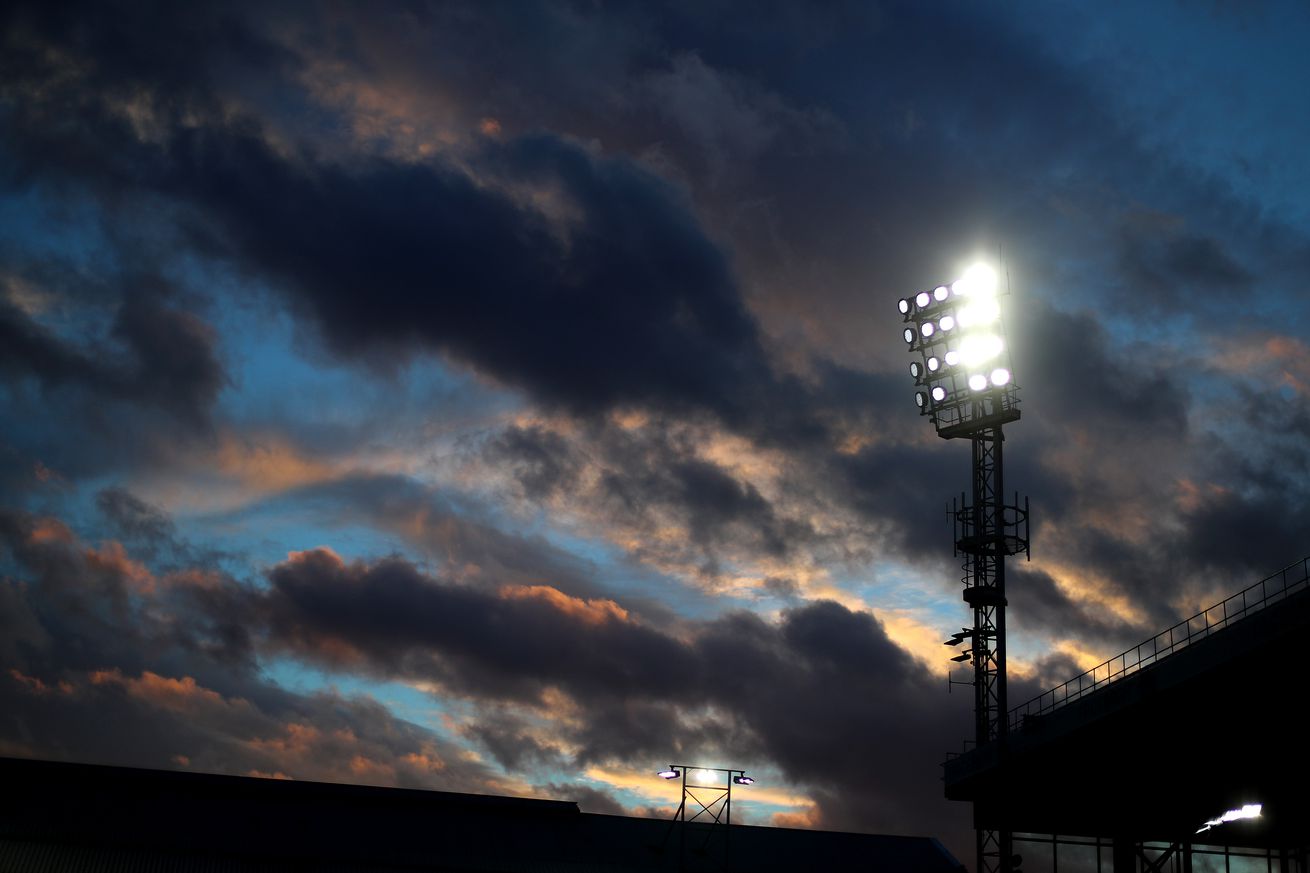 The sun sets over a stadium. Small broken clouds are illuminated in dark purples and reds, with some white cloud, fully bathed in sunlight, barely visible in the background. A single tower of stadium lights is in the frame, slightly off center, towards the right. It invokes the end of the day, a slight melancholy. Good night.