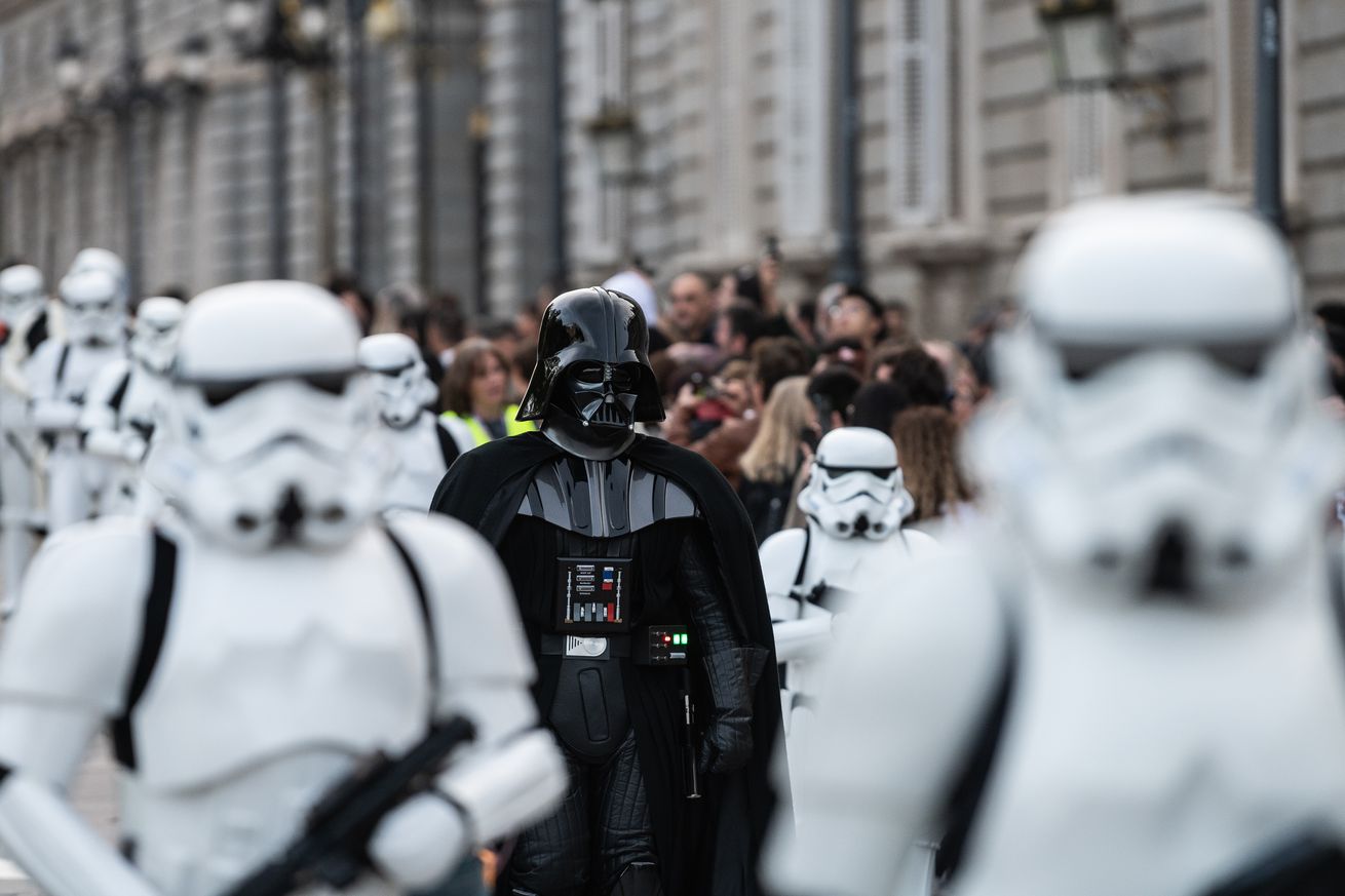 A man dressed as Darth Vader marching during a Star Wars parade surrounded by stormtroopers. 